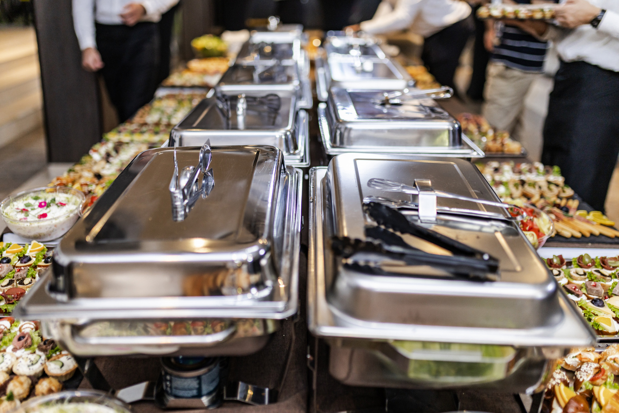 A sophisticated buffet is laid out, featuring an array of dishes including appetizers and desserts, with guests preparing to serve themselves. The atmosphere is lively and upscale.